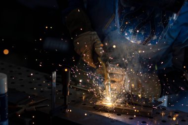 Kaynak maskeli, koruyucu giysili ve eldivenli bir adam atölyede çelik kaynaklıyor. Kıvılcımlı kaynak ateşi. Tehlikeli bir iş.