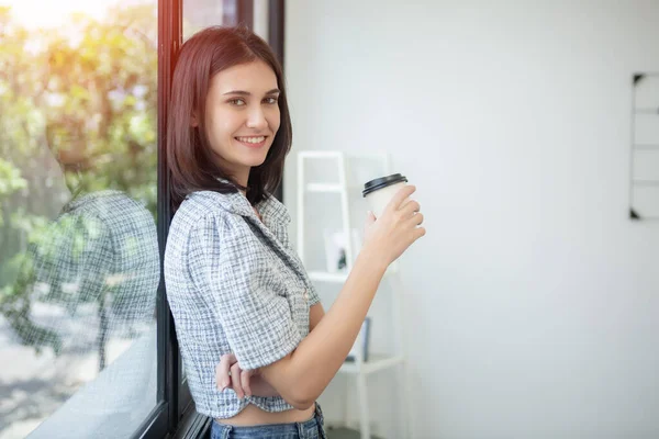 年轻美丽而快乐的女人拿着纸杯咖啡微笑望着窗外的办公室 亚洲女商人放松时间喝咖啡 — 图库照片
