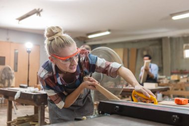 young carpenter woman wear uniform and goggles working use electronic saw cutting wood. craftsman profession in wood factory. woodworking industry.