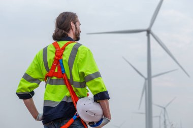 man engineer holding helmet safety wearing uniform standing  at wind turbine field renewable energy. technology protect environment reduce global warming problems. back view.