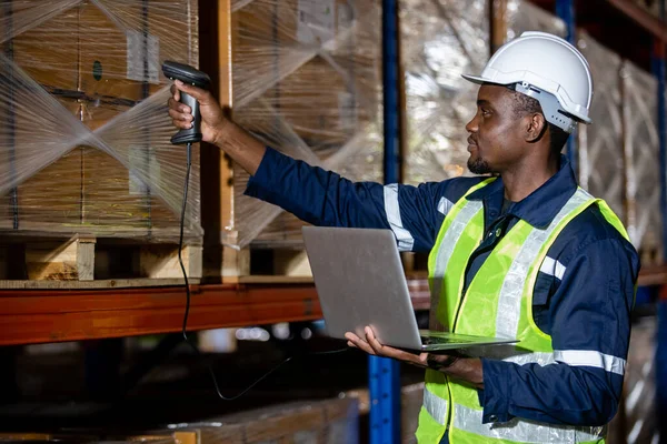 Black man professional worker wearing safety uniform and hard hat using laptop and scanning box inspect product on shelves in warehouse. Male worker is pointing finger inspecting product in factory.