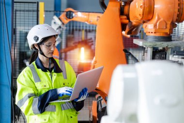 Dizüstü bilgisayar kullanan ve robot kolu otomatik kontrol eden kadın mühendis. Endüstriyel üretim sırasında robot otomasyonu. Fabrikada robotik test performansı.