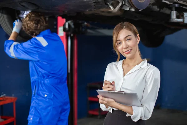 stock image Portrait of young woman holding clipboard standing at workplace with mechanic in garage background. Female employees taking notes on repair work and accounting work of car service company.