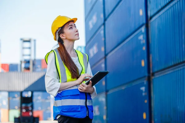 Female industrial engineer in uniform wearing safety hard hat using tablet checking containers loading. Area logistics import export and shipping cargo freight ship.