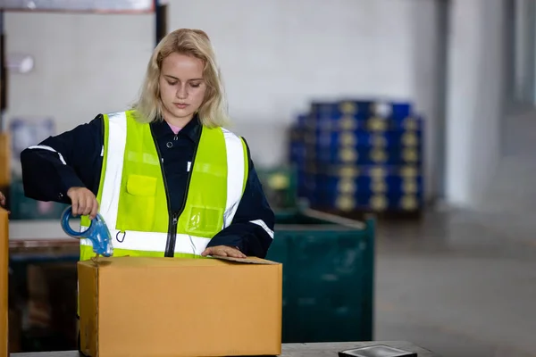 倉庫の段ボール箱製品に梱包テープを使用して安全制服を身に着けている女性ブロンドの髪のプロの労働者 — ストック写真