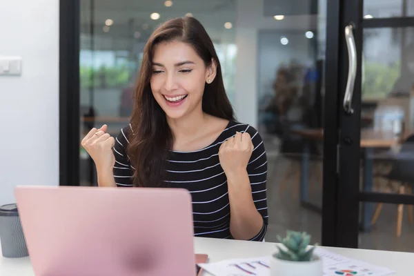 Happy young asian woman sitting at table with laptop showing hand success work at office. female work success looking at laptop.