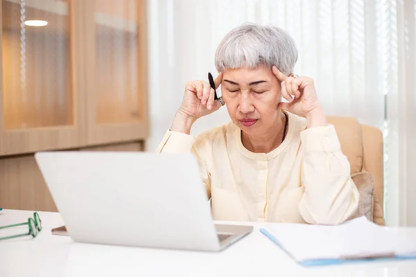 Stock image Asian senior business woman stress from hard work, Female manager headache sitting at table with laptop. Worried middle aged female health problems.