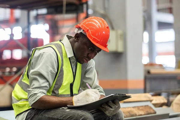 Happy male african american workers wear red helmet working take notes on paperwork clipboard sitting in factory. Heavy Industrial manufacturing workplace.