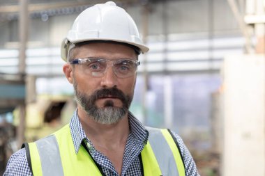 Kafkasyalı mühendis portresi beyaz şapka takıyor ve güvenlik üniforması giyiyor. Endüstriyel atölyede kameraya bakıyor. Teknik direktör imalatı. İşçiler büyük sanayi fabrikası.