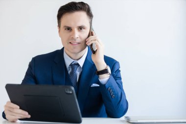 İş adamı telefonla konuşuyor. Ofis masasında dijital tableti tutan kameraya bakıyor. Yönetici şirkette telefon görüşmesi.