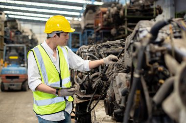 Asyalı mühendis üniforma ve miğfer giyerek depoda otomotiv parçaları kontrol ediyor. işyeri fabrikası endüstriyel ağır makine.