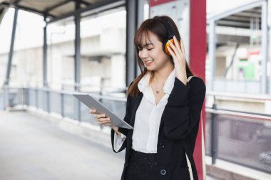 Siyah takım elbiseli çekici bir kadın elinde tablet, kulaklık takıyor gülümsüyor ve halka açık yerlerde kameraya bakıyor. Yaşam tarzı insan teknolojisi kavramı. Üniversitenin dışında bir kız öğrenci.