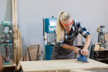 Genç marangoz beyaz kadın ahşap endüstrisinde elektrikli odun zımpara makinesi kullanıyor. Ahşap fabrikasında kadın zanaatkarlık mesleği.