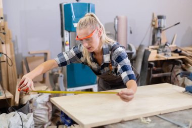 Kadın işçi ahşap endüstrisinde ahşap boyutlarında mezura kullanarak çalışıyor. Kadın marangoz, ahşap fabrikasında üniforma ve gözlük takar..