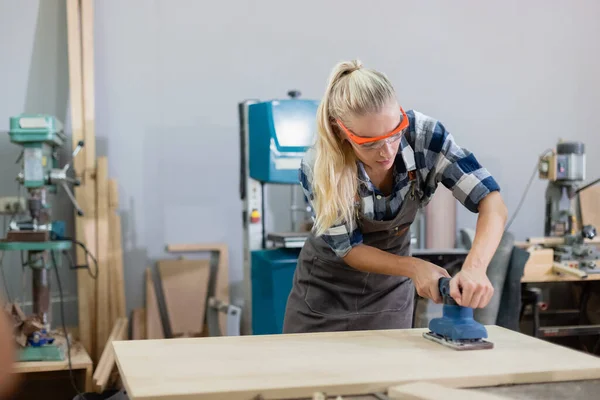 Stock image young carpenter caucasian woman using electric wood sander on wood at manufacturing wooden industry. Female craftsman profession in wood factory.