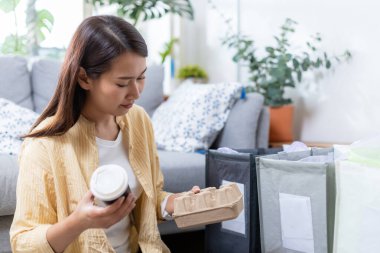 Asyalı genç bir kadın evde oturuyor kağıt ve plastik atıkları ayırıyor. Geri dönüşüm için çöp ayıklama. Küresel ısınmanın azaltılması. Çevre koruma kavramı.