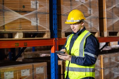 Depodaki raflarda dijital tablet denetim ürünü kullanarak güvenlik üniforması ve şapka takan profesyonel bir erkek işçi. İşçi, depolama lojistik fabrikasındaki stokları kontrol ediyor..