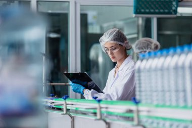 Kadın fabrika işçisi steril üniforma içecek suyu üretiyor. Şişe suyunu işlemek ve kontrol etmek için kadın fabrika hattı. Su içeceği üreten endüstriyel ürünler.