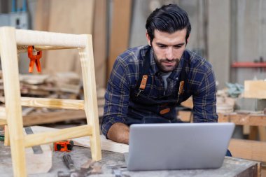 Dizüstü bilgisayarı olan genç bir marangoz çalışma alanında ahşap çizim yapıyor. Ahşap fabrikasında zanaatçılık mesleği