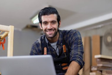 Dizüstü bilgisayarı olan genç bir marangoz çalışma alanında ahşap çizim yapıyor. Ahşap fabrikasında zanaatçılık mesleği