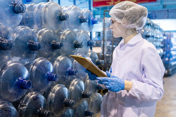 Женщина-работница держит планшет, чтобы проверить качество больших резервуаров питьевой воды перед процессом наполнения воды в пластиковые бутылки, чтобы принести потребителям. Бутылки с водой на производственной линии завода.