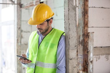 Erkek mühendis akıllı telefon kullanarak ev inşaatı yapıyor. Erkek mimar ev inşaatında duruyor. Ustabaşı iş yerinde.