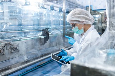 Kadın işçi, plastik şişelere su doldurup tüketicilere sunmadan önce kaliteli içme suyunu kontrol ediyor. Fabrika üretim hattında su şişeleri.