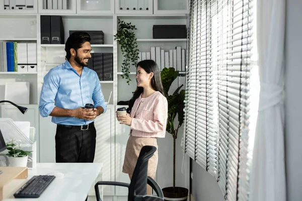 Man indian and asian woman holding coffee cup talking for work relax time standing in modern office. Asian business people meeting talking brainstorming together in office Teamwork concept.