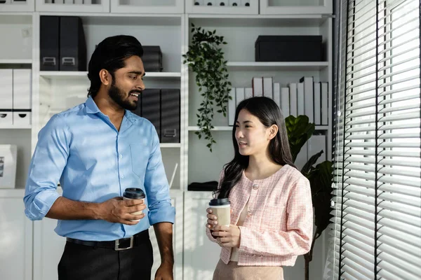 Man indian and asian woman holding coffee cup talking for work relax time standing in modern office. Asian business people meeting talking brainstorming together in office Teamwork concept.