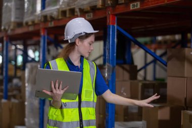 Depodaki raflarda dizüstü bilgisayar kullanarak güvenlik üniforması ve şapka takan kadın işçi. Kadın işçi fabrikada hisse senedi teftişi yapıyor..