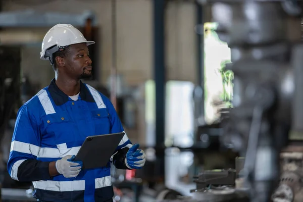 Male African American workers wearing uniform safety and hardhat using tablet working at machine in factory Industrial. Engineering worker man work machine lathe metal. Heavy industry concept.