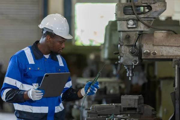 Male African American workers wearing uniform safety and hardhat using tablet working at machine in factory Industrial. Engineering worker man work machine lathe metal. Heavy industry concept.
