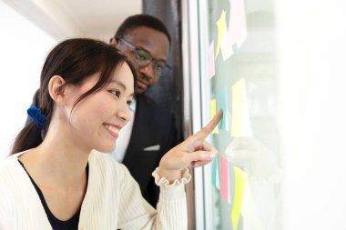 Çok ırklı iş adamları toplantısı, cam tahtadaki notlara işaret ediyor. İşletme ekibi beyin fırtınası fikri ofiste toplanıyor. Şirket pazarlama stratejisi projesi planlanıyor.