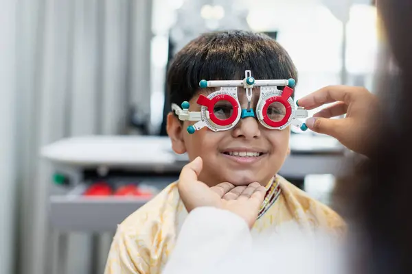 クリニックで視力の現代眼科機器を調べる女性眼科の専門家を持つ幸せなインドの子供の少年 患者の男性検診アイリスは眼科病院を診察する 眼鏡を測定する ストック画像