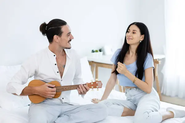 happy couple playing ukulele on bed in bedroom. man and woman enjoying playing music creates happy activities in family. young couple fun happiness romantic with instrument.