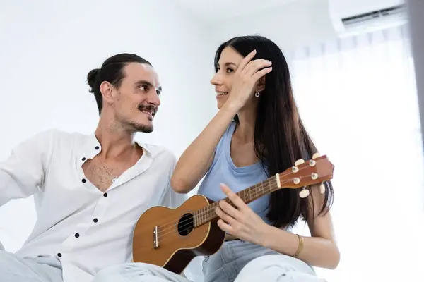 happy couple playing ukulele on bed in bedroom. man and woman enjoying playing music creates happy activities in family. young couple fun happiness romantic with instrument.