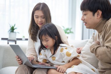 Mutlu Asyalı aile babası anne ve kızı evdeki oturma odasındaki kanepede oturan dizüstü bilgisayarla eğleniyorlar. Ebeveynler çocuk kızlara internet tarayıcılığı öğrenme becerisi öğretiyor.