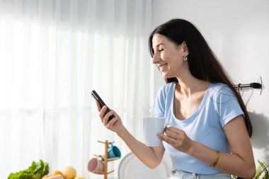 Kafkasyalı bir kadın sabah kahvesi içiyor. Modern mutfak tezgahında oturmuş akıllı telefon okuyor. Kadın kahvaltısı sosyal medya sohbeti.