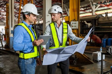 Tren yolu teknisyeni üniformalı güvenlik kaskı takıyor. Tablet ve taslak kontrol planı demiryolu istasyonunda duruyor. Fabrikada lokomotif tamircisi.