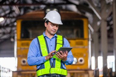 Demiryolu teknisyeni üniformalı ve kasklı bir adam tren istasyonunda tablet tutuyor. Fabrikada lokomotif tamircisi.