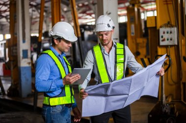 Tren yolu teknisyeni üniformalı güvenlik kaskı takıyor. Tablet ve taslak kontrol planı demiryolu istasyonunda duruyor. Fabrikada lokomotif tamircisi.