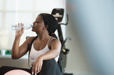 Spor salonunda egzersiz yaptıktan sonra şişede spor giyinen sağlıklı, güçlü, Afro-Amerikan kadın sporcu kıyafeti suyu içmek vücudu canlandırır. Rahatla ve egzersiz konsepti.