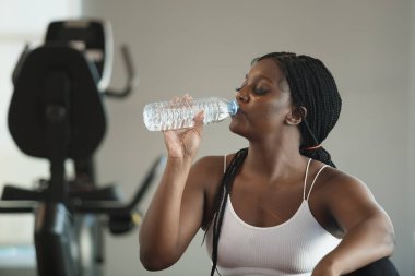 Spor salonunda egzersiz yaptıktan sonra şişede spor giyinen sağlıklı, güçlü, Afro-Amerikan kadın sporcu kıyafeti suyu içmek vücudu canlandırır. Rahatla ve egzersiz konsepti.