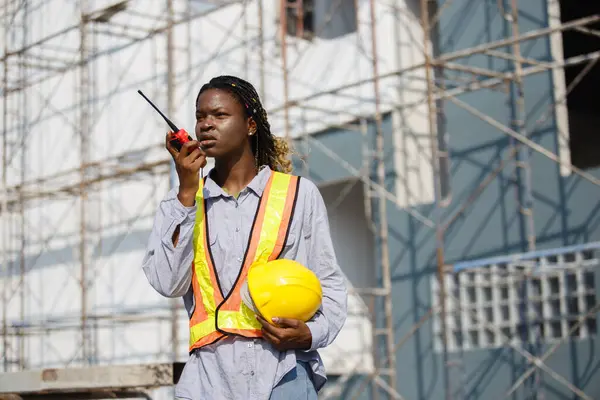 African American women general employee operational level wear yellow hard hat holding talkie walkie at village construction site. Construction worker talking radio.
