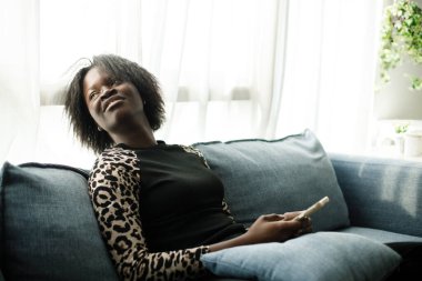 Genç Afro-Amerikan kadın modern ev oturma odasındaki koltukta akıllı telefon kullanıyor. Konuşmak ve internetten alışveriş yapmak için cep telefonu kullanmak. İnternet üzerinden film izlemek.