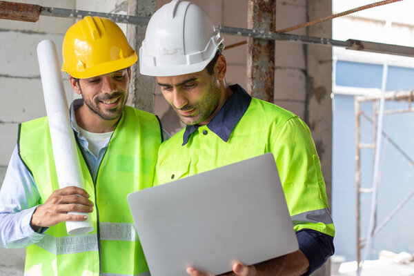 Профессиональная команда инженеров-строителей, двое инженеров носят униформу, держат ноутбук и смотрят на камеру, стоящую на стройке. Руководитель архитектурного бюро.