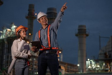 Asyalı ekip elemanı ve kadın mühendis gece yarısı petrolle çalışan elektrik santralinin önünde planlarla çalışıyorlar. İşçiler kule inşaatında çalışan üniformalı miğfer giyerler..