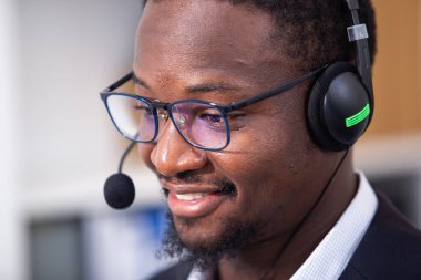 Kulaklık takan Afro-Amerikan arkadaş canlısı bir adam çağrı merkezi çalışanı müşteri destek operatörü ofisteki bilgisayarı kullanıyor. İş çevrimiçi müşteri hizmetleri merkezi.