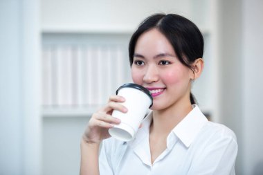 Genç, güzel, neşeli bir kadın elinde kağıt bardakla pencereden bakan gülümseyen bir kadın. Asyalı iş kadını kahve içerek rahatlıyor..