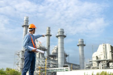 Asyalı işçi fabrikası planlarını tutuyor. İnşaat planının elektrik santralini işaret ediyor. Enerji petrokimya endüstrisinde güvenlik üniforması giyen mühendis..